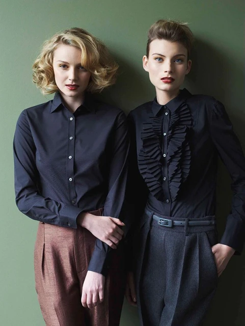 Two fair-skinned women pose indoors against a green wall. They both wear long-sleeve shirts and high-waisted pants in a fashion editorial.