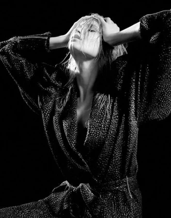 Black and white portrait of a model in a patterned wrap dress with hands on head, set against a dark, high-contrast background.