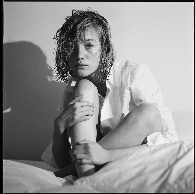Black and white portrait of a young woman with wet hair, wearing an unbuttoned white shirt, sitting on a white bed, lit by side lighting.