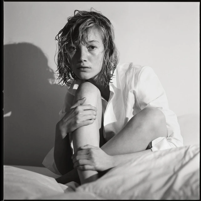 Black and white portrait of a young woman with wet hair, wearing an unbuttoned white shirt, sitting on a white bed, lit by side lighting.