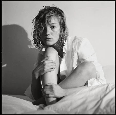 Black and white portrait of a young woman with wet hair, wearing an unbuttoned white shirt, sitting on a white bed, lit by side lighting.