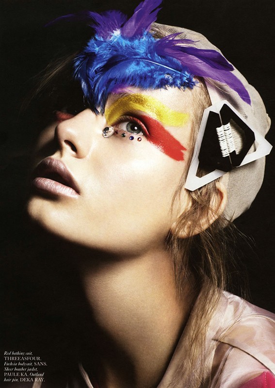 Model wearing a headpiece with colorful feathers and abstract makeup posing in a beauty and fashion editorial shot.