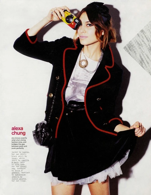 Fashion editorial shot of a woman holding a camera, wearing a black jacket with red trim, a black skirt, and multiple necklaces.