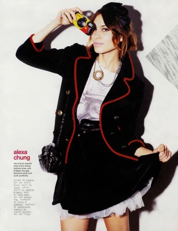 Fashion editorial shot of a woman holding a camera, wearing a black jacket with red trim, a black skirt, and multiple necklaces.