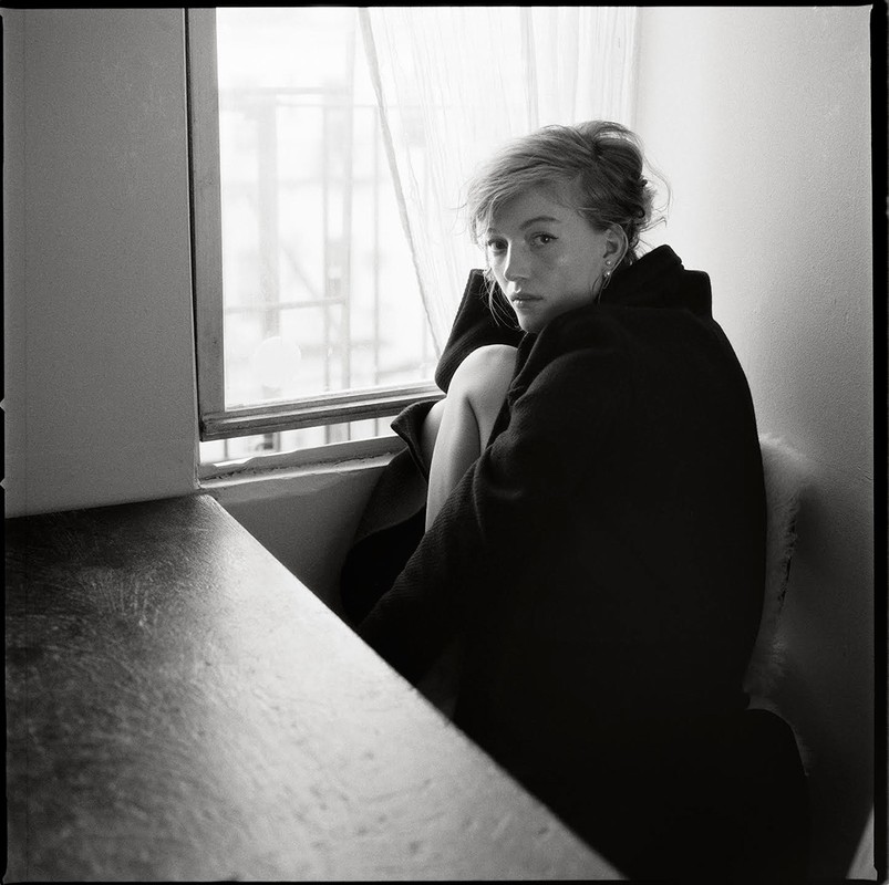 Black and white portrait of a woman with tousled hair seated by a window, wearing a dark coat.