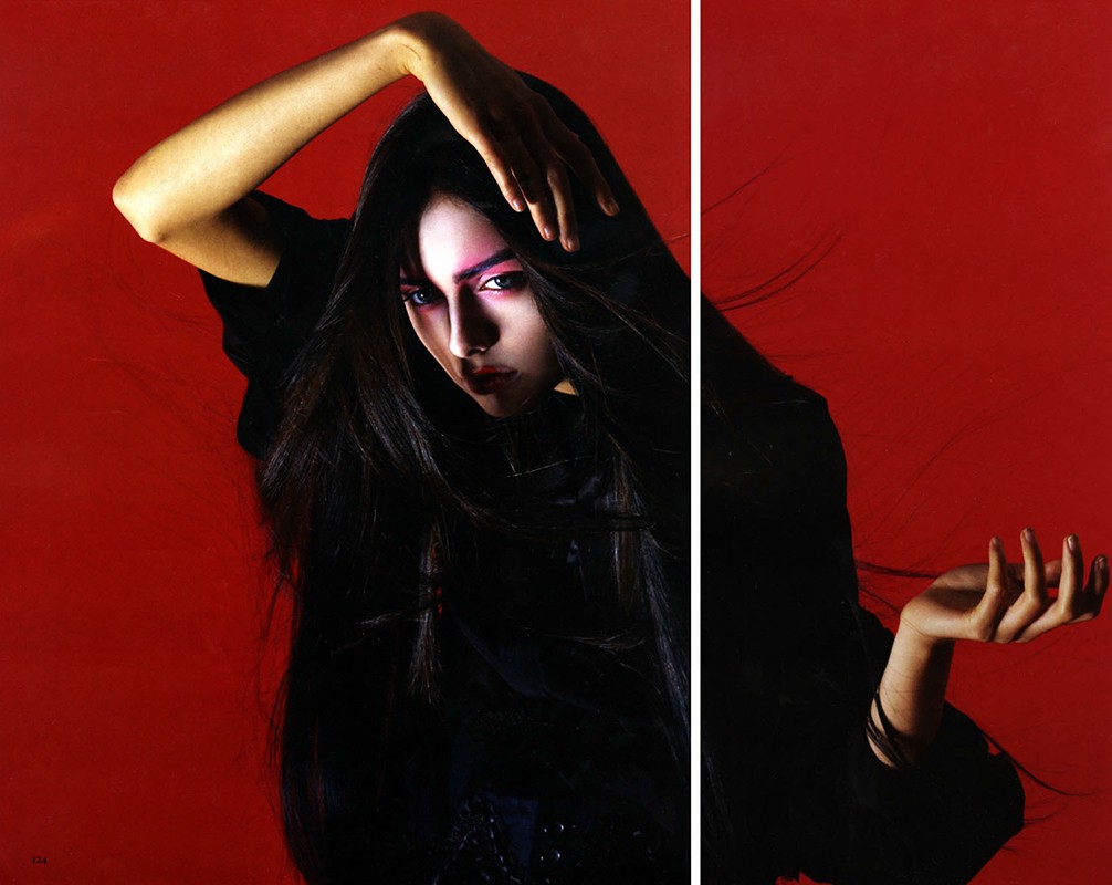 Portrait of a woman with long black hair, wearing black clothing and pink eye shadow, against a red background in a studio setting.