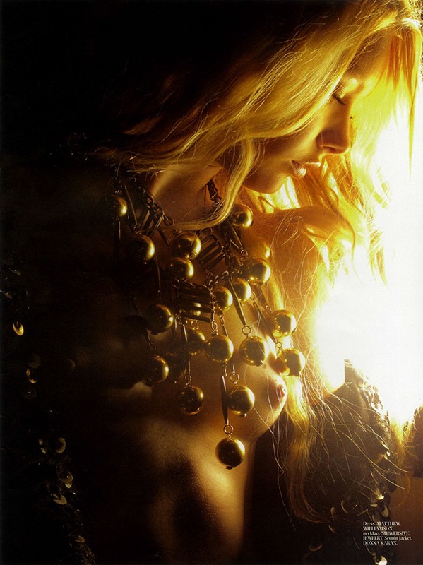 Blonde woman poses with a golden necklace in a low-key lighting setup. The soft, warm light emphasizes the golden jewelry and her hair.