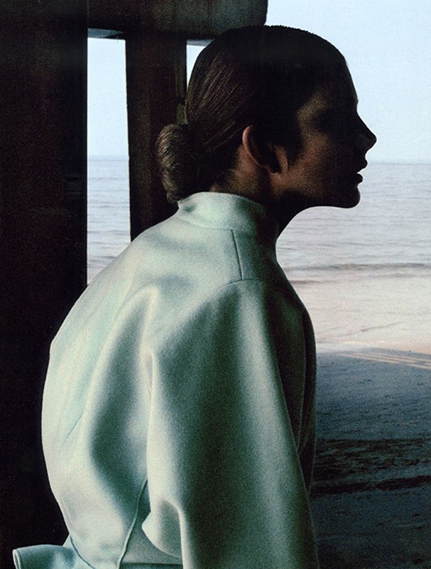 Profile view of an adult female in a light green high-neck jacket at a beach. She has her hair in a bun, and is looking out at the ocean.
