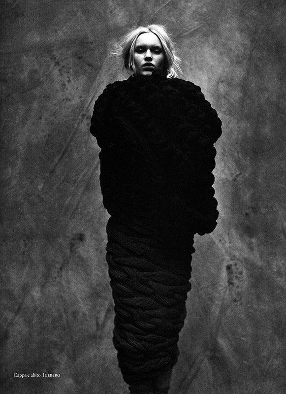 Monochrome fashion portrait of a woman cocooned in a large, textured black garment against a mottled grey backdrop in a studio shot.