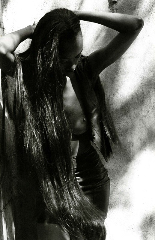 Monochrome portrait of a slender, topless adult woman with long, dark hair posed in front of a mottled wall with dappled sunlight and shadows.