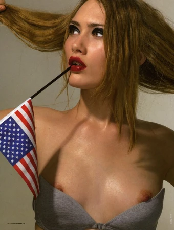 Close-up portrait of a woman pulling her long hair, holding a small American flag in her mouth against a grey background.