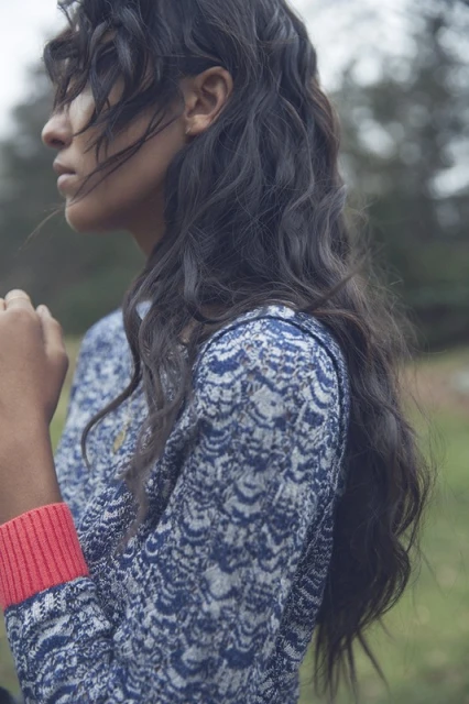 Portrait of an adult woman with dark wavy hair, wearing a blue and white patterned cardigan in an outdoor, grassy environment.