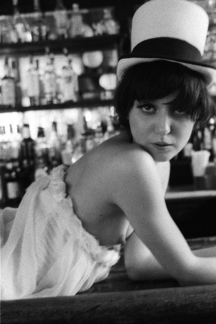 Monochrome portrait of a woman in a top hat leaning on a bar counter, with a background of bottles and glasses on shelves.