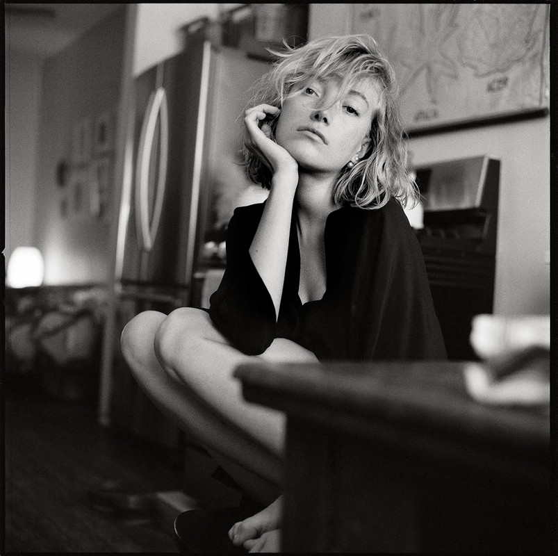 Black and white portrait of a woman with blonde hair sitting indoors near a stainless steel refrigerator and a dark wooden table.