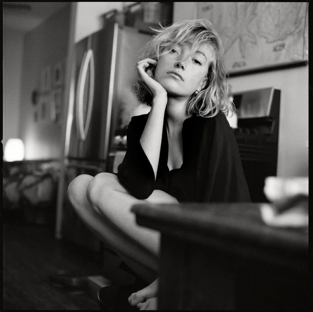 Black and white portrait of a woman with blonde hair sitting indoors near a stainless steel refrigerator and a dark wooden table.