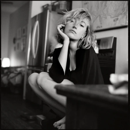 Black and white portrait of a woman with blonde hair sitting indoors near a stainless steel refrigerator and a dark wooden table.