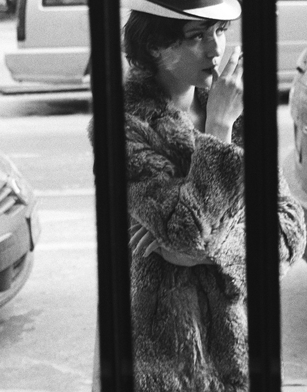 Monochrome portrait of a woman in a fur coat and hat, sipping from a cup, framed by vertical bars with blurry cars in the background.