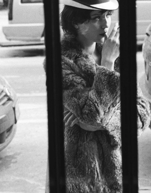 Monochrome portrait of a woman in a fur coat and hat, sipping from a cup, framed by vertical bars with blurry cars in the background.