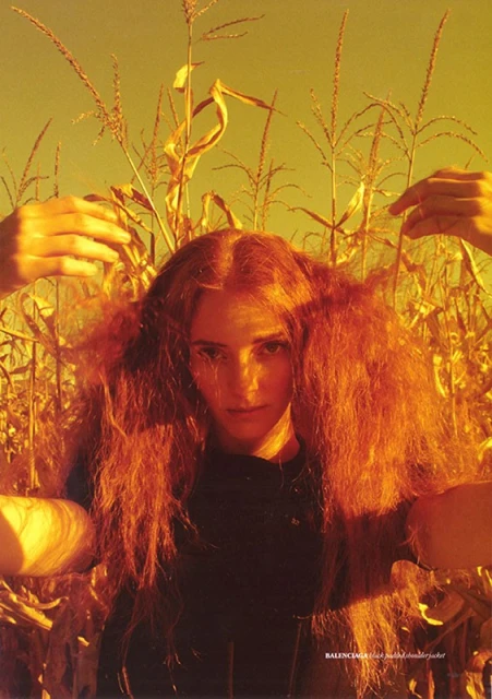 Fashion editorial portrait of a redhead woman in a dark top standing in a cornfield, with hands framing her face.