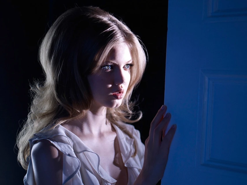 Blonde woman with long, wavy hair, fair skin, and light eyes stands near a blue door in a dimly lit indoor setting, close-up shot.