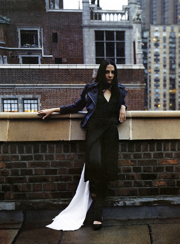 Woman in black outfit with long white train posing on rooftop with city buildings in the background in medium shot, fashion photography.