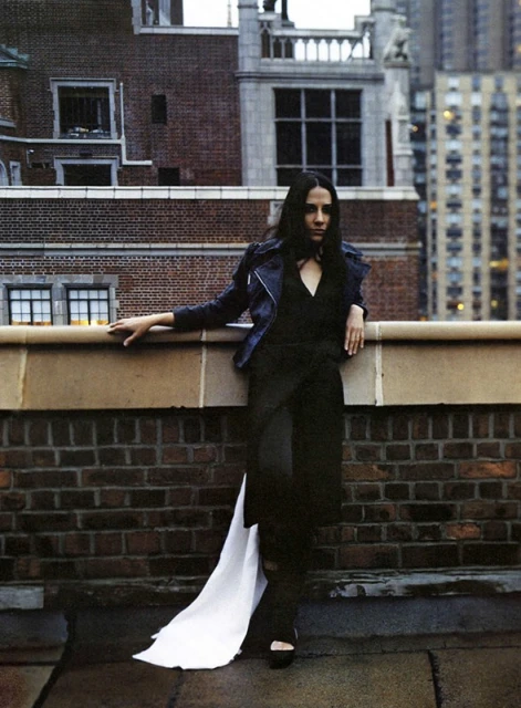 Woman in black outfit with long white train posing on rooftop with city buildings in the background in medium shot, fashion photography.