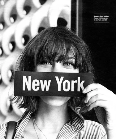 Black-and-white shot of a woman holding a 'New York' sign over her face, focusing on her eyes and shaggy hair with a patterned background.