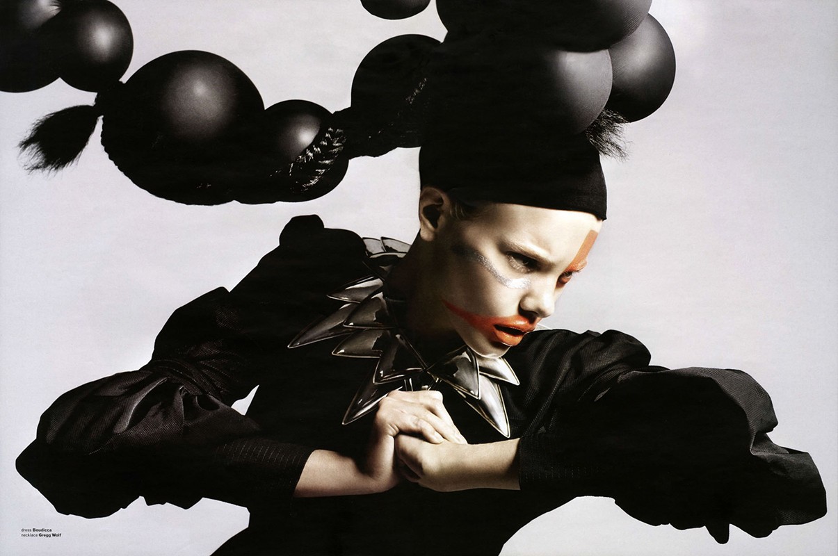 Fashionable model with dark makeup and spiked collar, black top, black hair styled into round shapes against a stark white background.