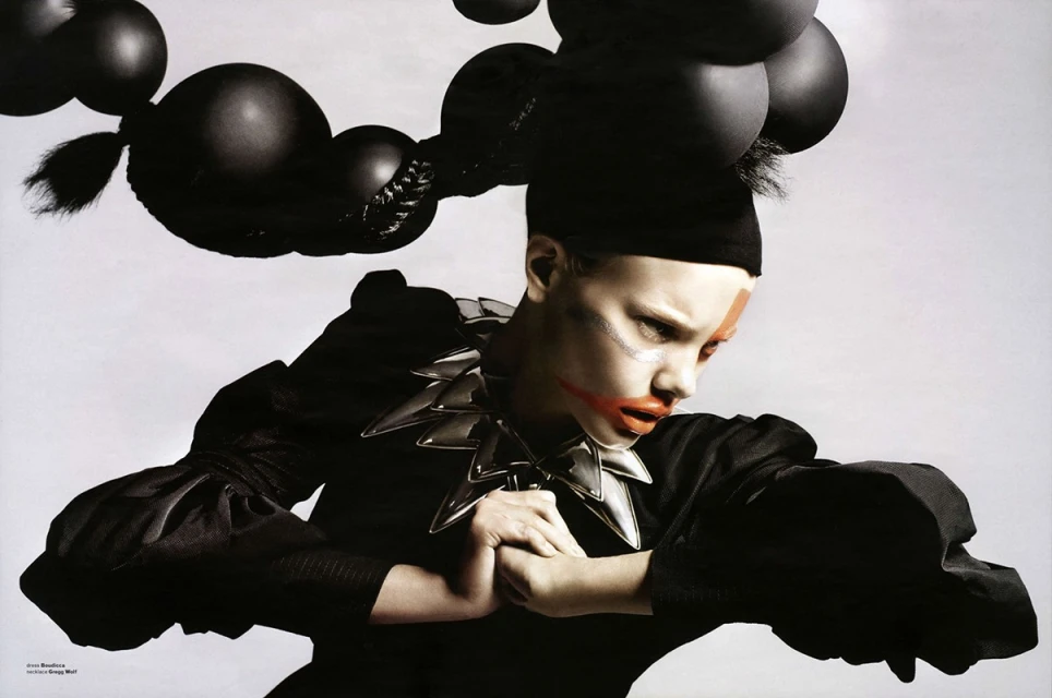 Fashionable model with dark makeup and spiked collar, black top, black hair styled into round shapes against a stark white background.