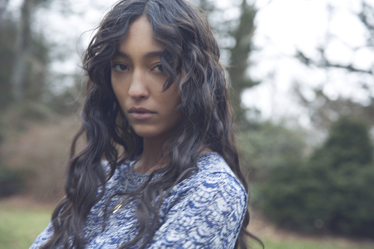 Portrait of a young adult female with long dark wavy hair, wearing a printed top, outdoors in a blurry natural setting with trees.