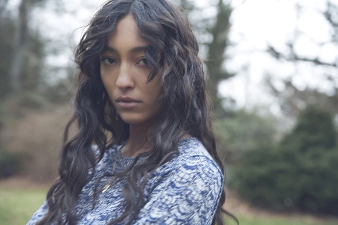 Portrait of a young adult female with long dark wavy hair, wearing a printed top, outdoors in a blurry natural setting with trees.