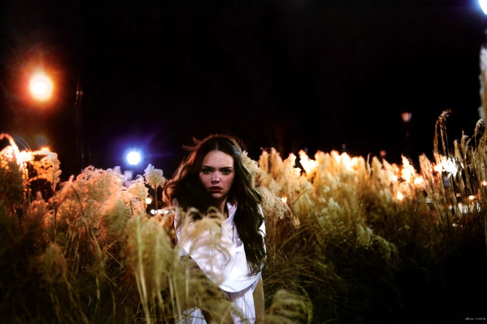Young woman stands among tall grass at night, illuminated by distant lights; she is wearing a white top and looking at the camera.
