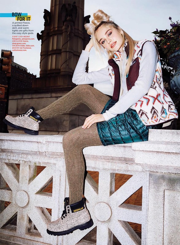 A fashionable young woman with blonde hair sits on a ledge wearing a printed fleece vest, long sleeve top, green shorts, patterned tights, and boots.