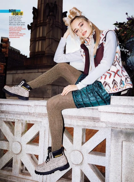 A fashionable young woman with blonde hair sits on a ledge wearing a printed fleece vest, long sleeve top, green shorts, patterned tights, and boots.