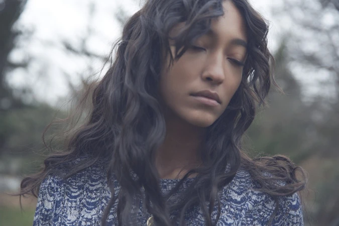 Close-up portrait of a woman with long dark wavy hair and closed eyes wearing a patterned blue knit top outdoors.