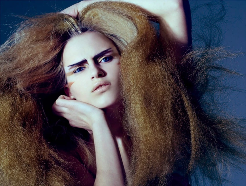 Dramatic beauty portrait featuring a woman with exaggerated hair, bold makeup, and striking blue eyes against a dark background.