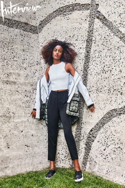 Young woman poses in front of a textured wall wearing a light top, dark pants, an unbuttoned jacket, and sneakers on green grass.