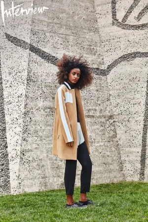 Woman with voluminous curly hair wearing a camel coat standing against a textured concrete wall