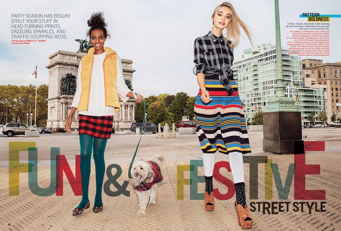 Two women model street style fashion in an urban setting with a dog. One wears plaid, the other a vest and skirt. Buildings in the background.