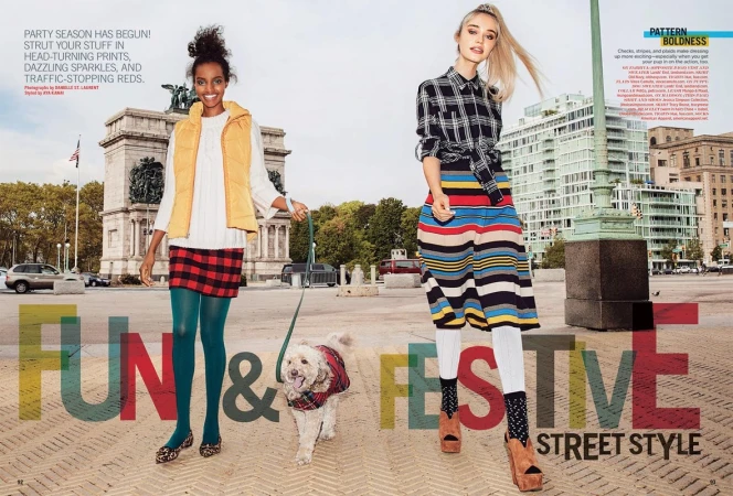 Two women model street style fashion in an urban setting with a dog. One wears plaid, the other a vest and skirt. Buildings in the background.