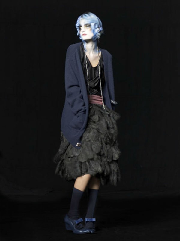 Full-length shot of a fashion model with blue hair and vintage-style clothing posing against a dark backdrop in a studio setting.
