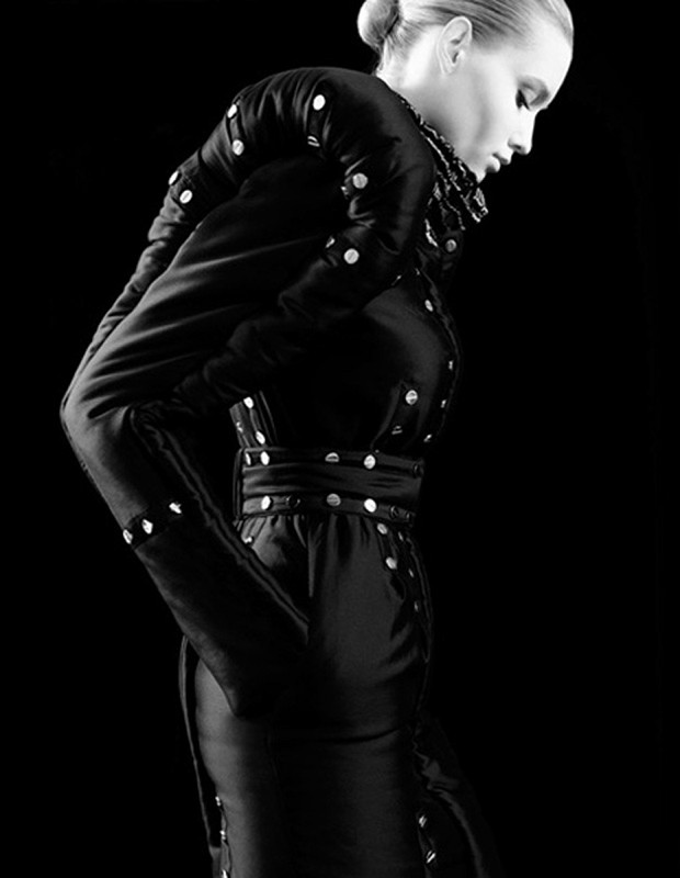 Fashion shot of a slender adult female model in an elegant black dress with white accents against a stark black studio background. Side view.