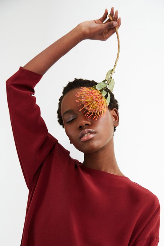 Portrait of an African American woman in red shirt with flower over her eye in a light studio, eyes closed, neutral facial expression.