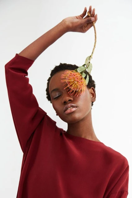 Portrait of an African American woman in red shirt with flower over her eye in a light studio, eyes closed, neutral facial expression.