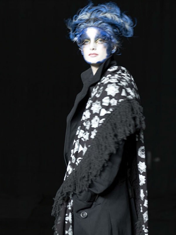 Woman with blue and white hair, wearing black coat and floral shawl in a dark studio, highlighted with dramatic makeup and artistic styling.