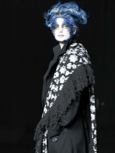 Woman with blue and white hair, wearing black coat and floral shawl in a dark studio, highlighted with dramatic makeup and artistic styling.