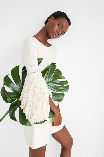 Dark-skinned woman in a white dress poses with a large monstera leaf against a white background in a fashion-style studio shot.