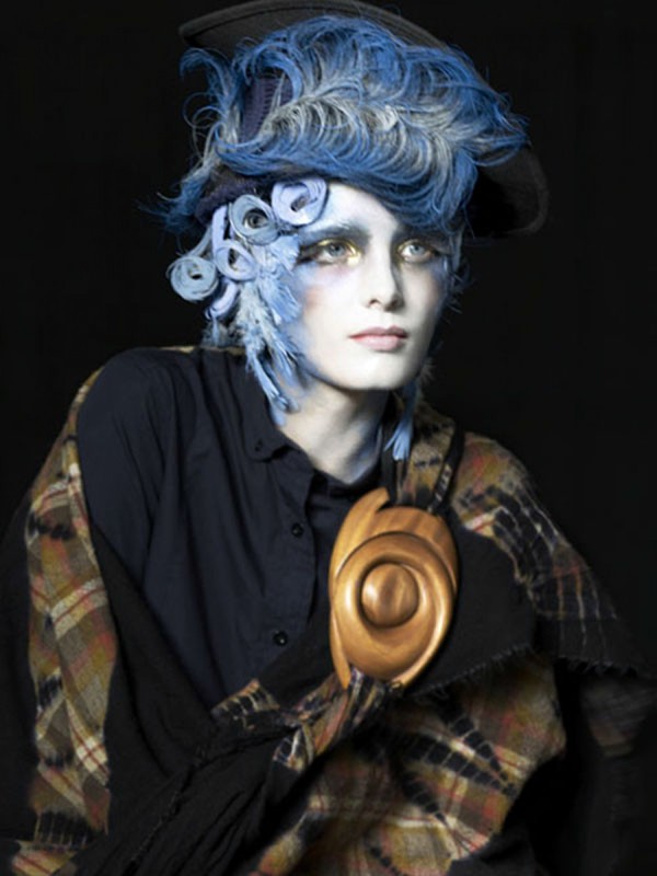Dramatic beauty portrait: person with stylized blue hair, theatrical makeup, plaid shawl, and an ornate wooden necklace against black.