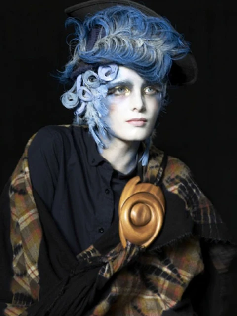 Dramatic beauty portrait: person with stylized blue hair, theatrical makeup, plaid shawl, and an ornate wooden necklace against black.