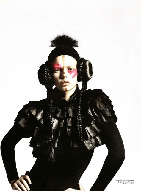 Avant-garde fashion portrait of a person with pink face paint, wearing a black knit hat and an elaborate, tiered leather cape on a white background.